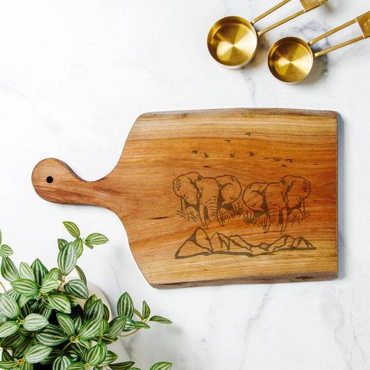 Wooden cutting board with engraved design on a white surface with gold measuring spoons and a plant.